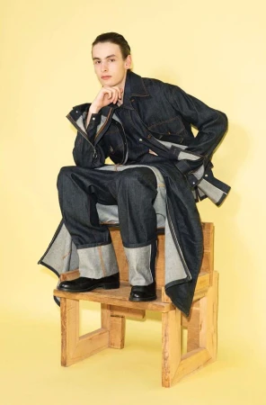 Young male model in dark denim jacket and cuffed trousers sitting on a rustic wooden chair against a pale yellow background.