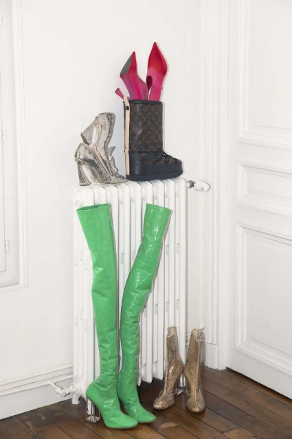 Several pairs of stylish women's boots and high heels of different designs and colors sit atop a radiator in an indoor setting.