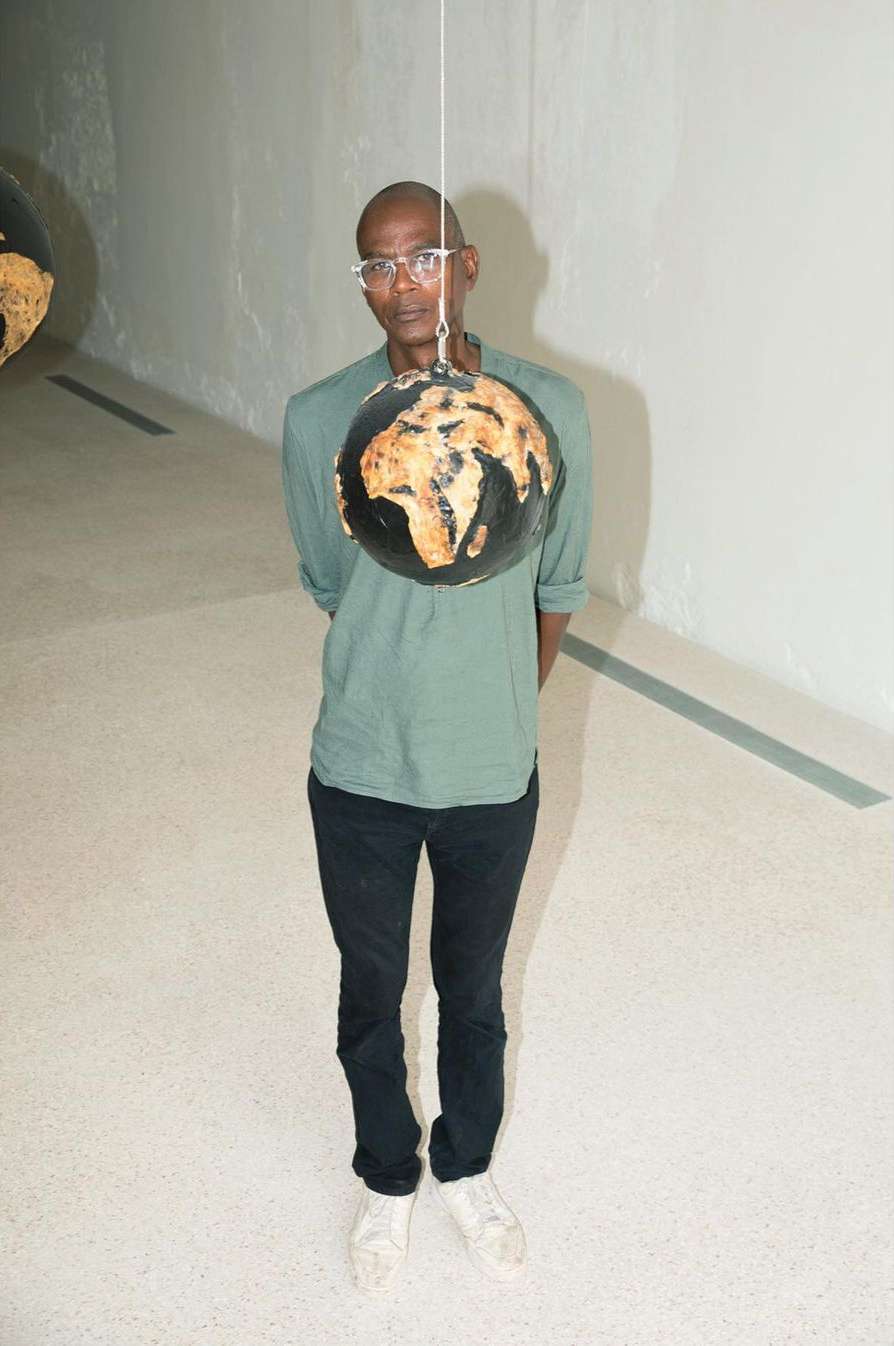 Full shot of adult Black man in eyeglasses standing indoors with stringed globe. He wears a green long sleeve shirt, dark jeans and white shoes.