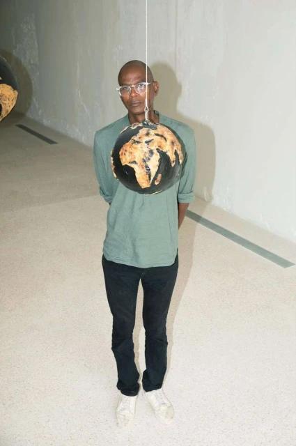 Full shot of adult Black man in eyeglasses standing indoors with stringed globe. He wears a green long sleeve shirt, dark jeans and white shoes.