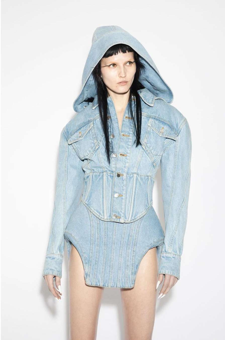 A young adult fashion model in a light-blue denim hooded jacket and matching denim bottoms poses in a brightly lit photo studio.