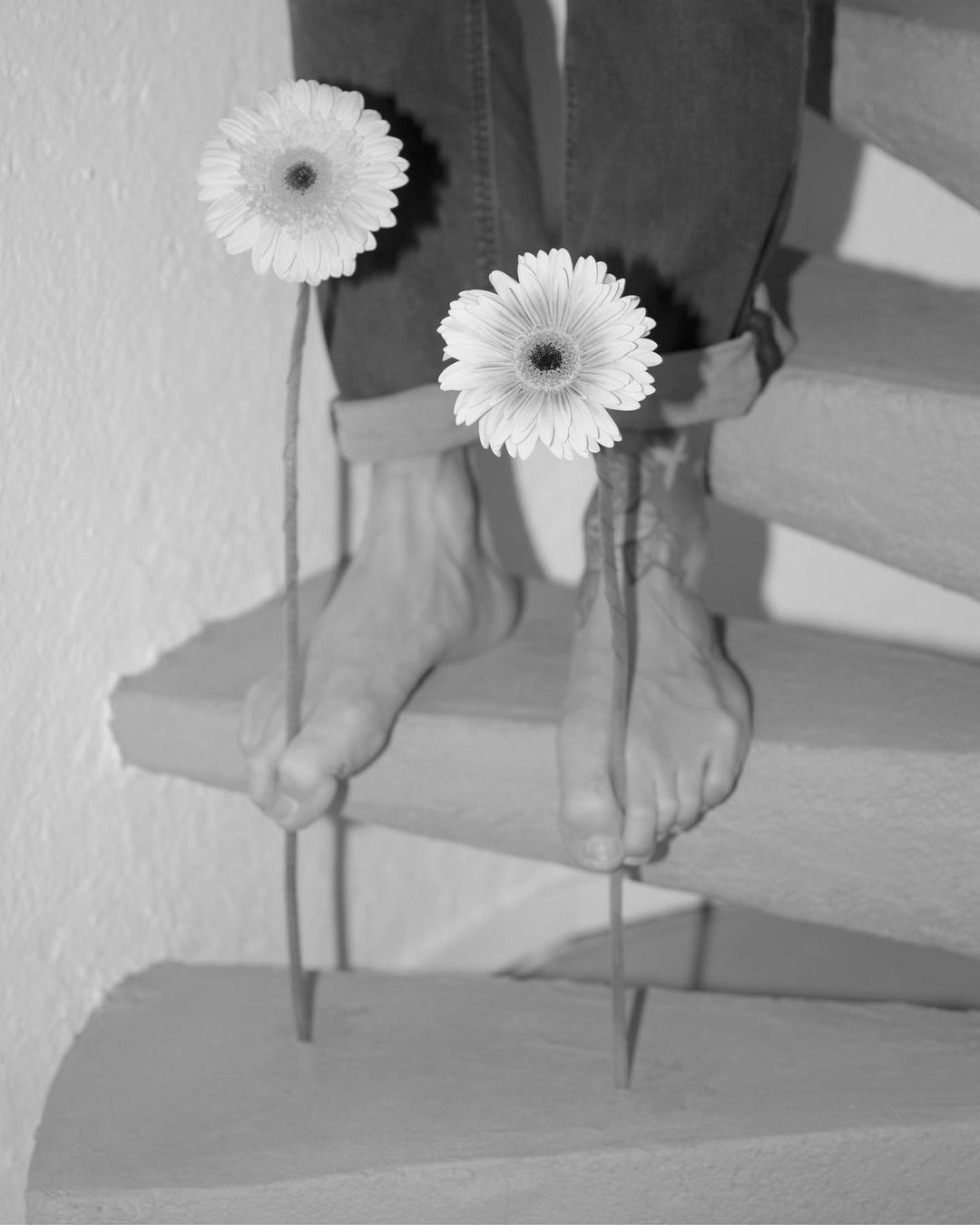 Black and white image of a person's feet standing on a staircase with two daisy flowers in between, creating an artistic monochrome composition.