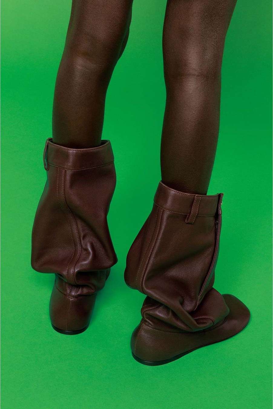 Close-up of a person's lower legs and feet wearing dark brown leather slouchy flat boots against a bright green background.