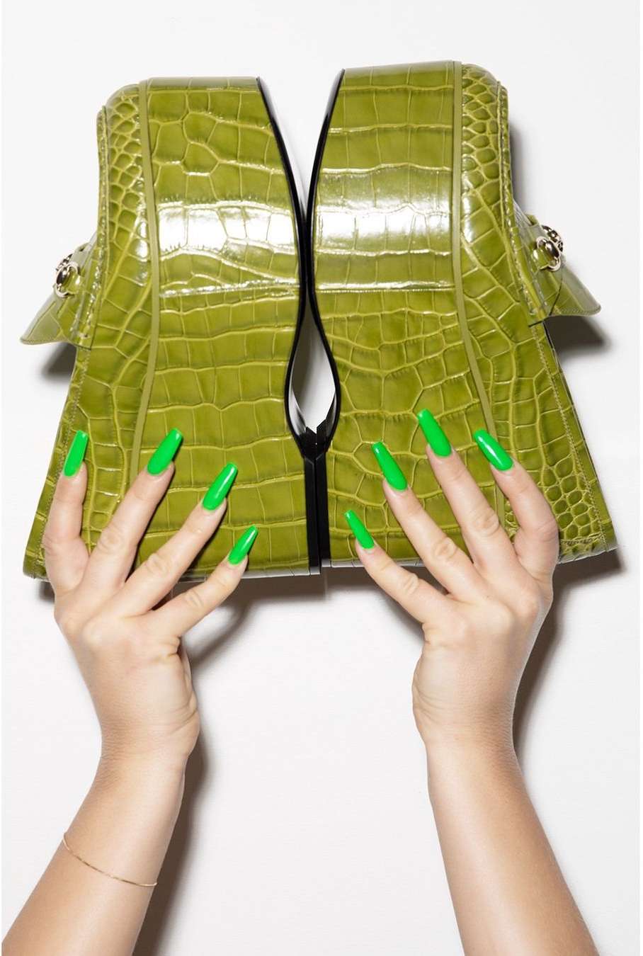 A pair of platform loafers in green crocodile leather are held up by hands with matching green manicured nails, shot in a photo studio.