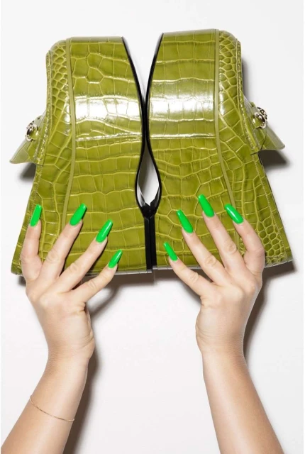 A pair of platform loafers in green crocodile leather are held up by hands with matching green manicured nails, shot in a photo studio.