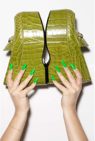 A pair of platform loafers in green crocodile leather are held up by hands with matching green manicured nails, shot in a photo studio.