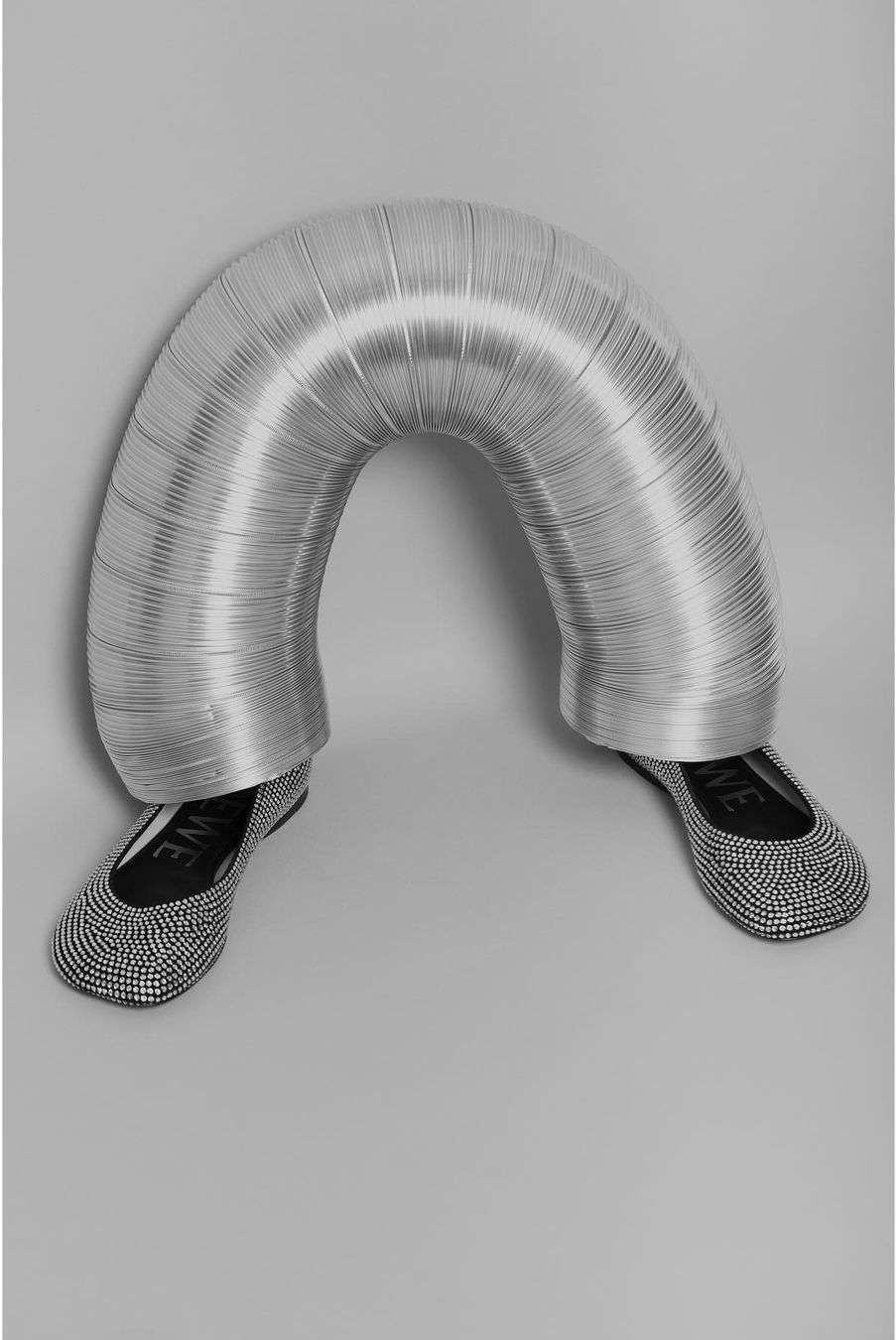 Eye-level shot featuring a pair of ballet flats with a person partially visible under a corrugated metal pipe in a grey studio.