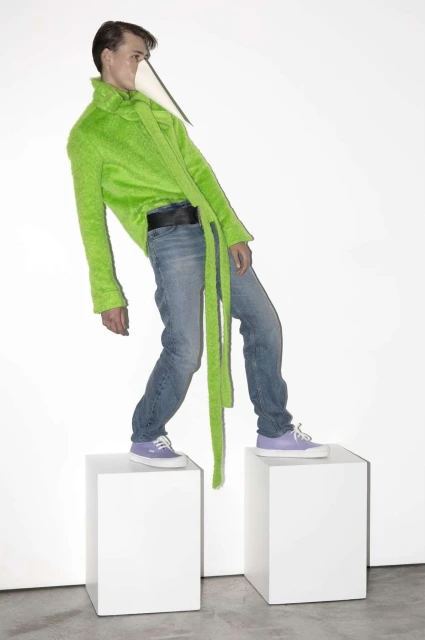 Adult male fashion model stands on white blocks wearing a lime-green jacket, blue jeans, and lilac sneakers against a white backdrop.