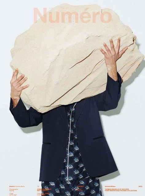 Magazine cover with a person holding a large rock over their head, wearing a blazer and patterned pajamas against a white background.