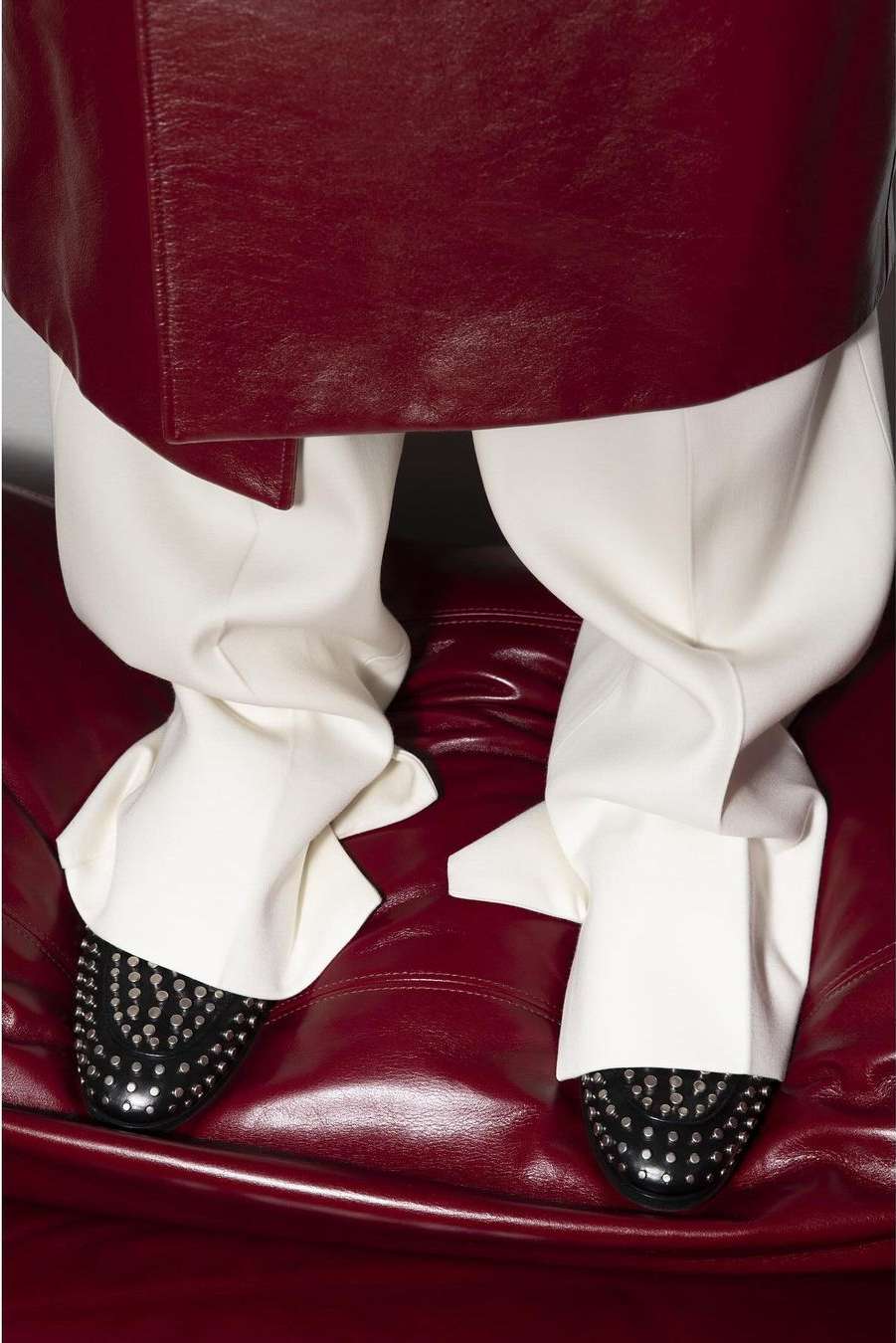 Close-up of lower body of a person wearing a burgundy coat, white trousers, and black studded dress shoes, seated on a burgundy couch.