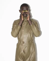 Black man wearing an olive green leather jumpsuit holding grapes over his eyes against a plain white background