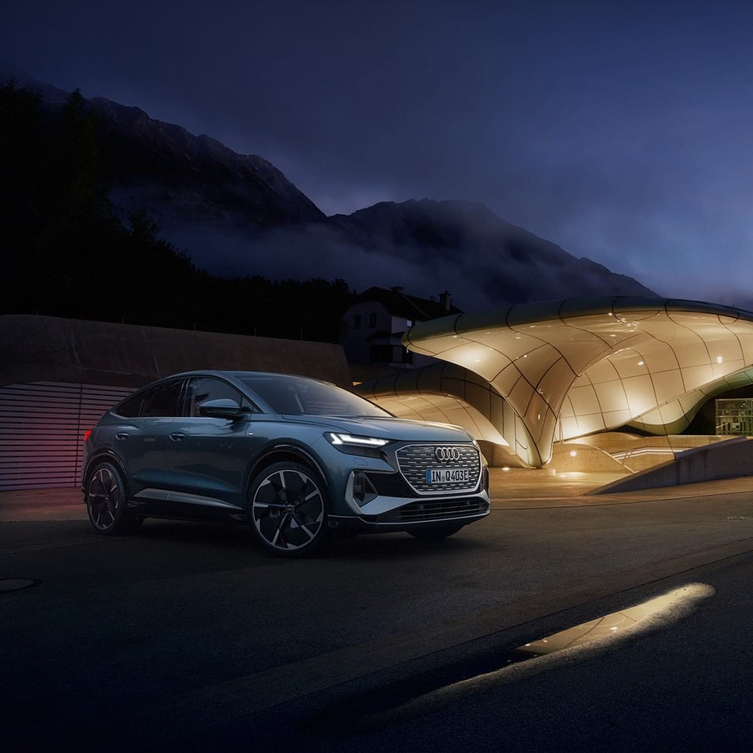 An Audi Q4 e-tron electric vehicle parked at night with a futuristic building in the background and mountain range in the distance.