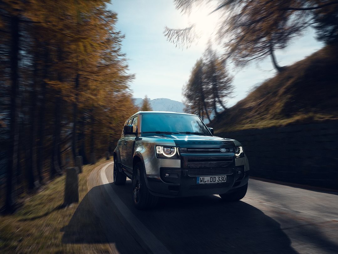 Silver Land Rover Defender driving on a rural road surrounded by autumn trees and mountains under a blue sky in motion blur.