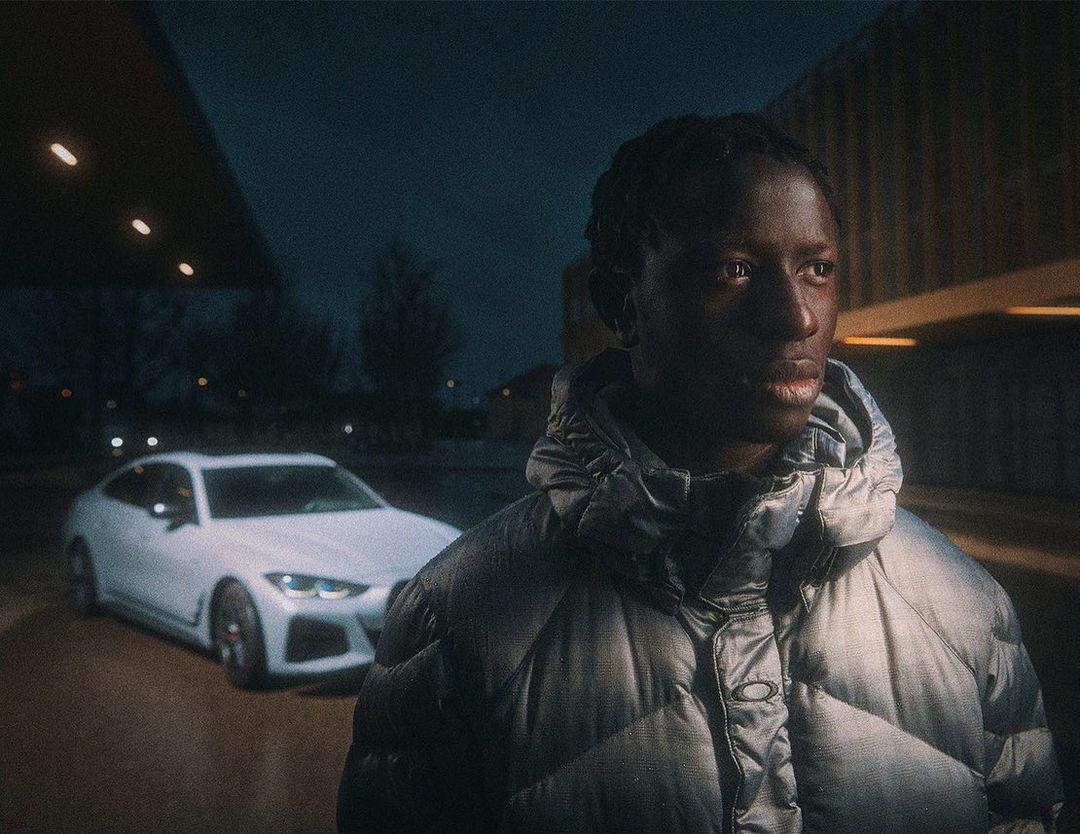 Adult male poses outdoors in silver puffer jacket next to modern white car at dusk with modern architecture in background.