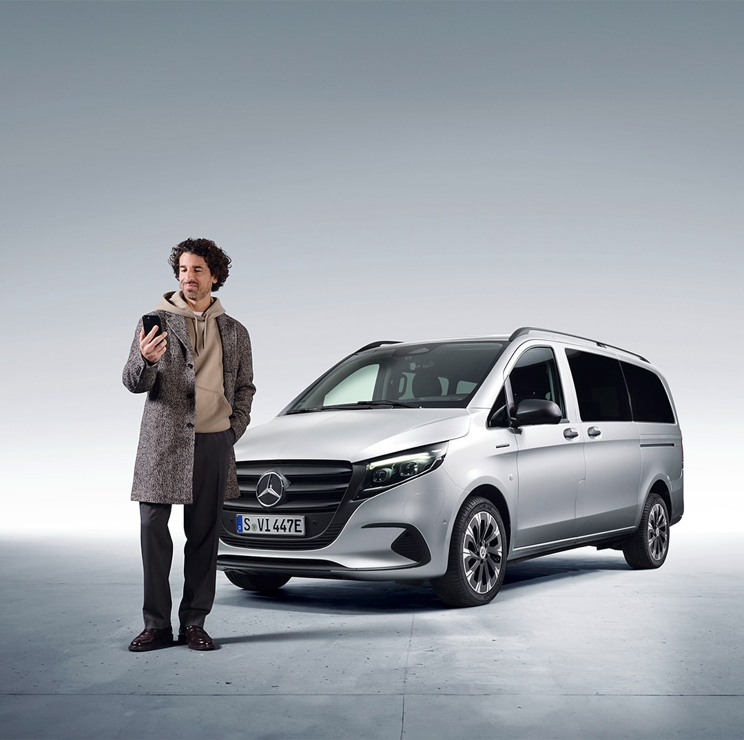 Adult male looks at his smartphone next to a silver Mercedes-Benz EV van in a photo studio with soft, diffused lighting.