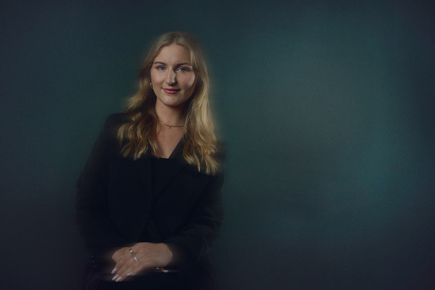 Portrait of a young blonde woman in a dark blazer against a blurred teal background, showcasing a professional and elegant headshot.