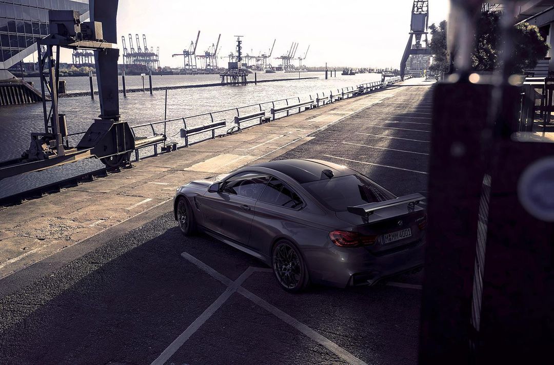 Sleek grey BMW coupe parked on a paved road near a waterfront, with shipping cranes visible in the background on a bright day.