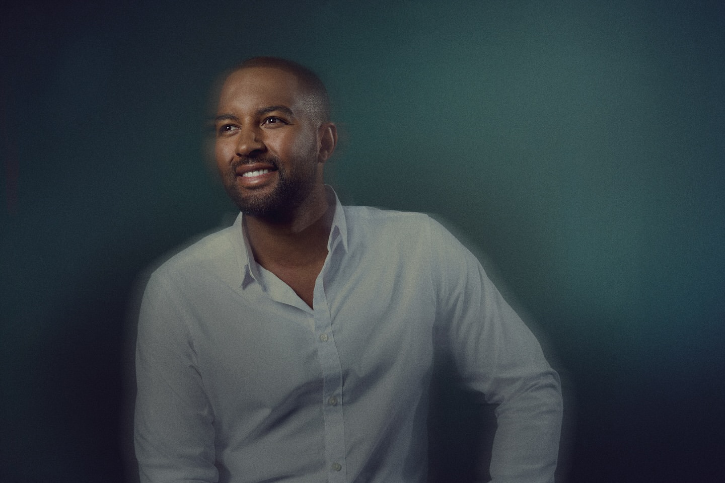 Portrait of a smiling adult male with short dark hair in a white button-down shirt against a teal studio backdrop in eye-level shot.
