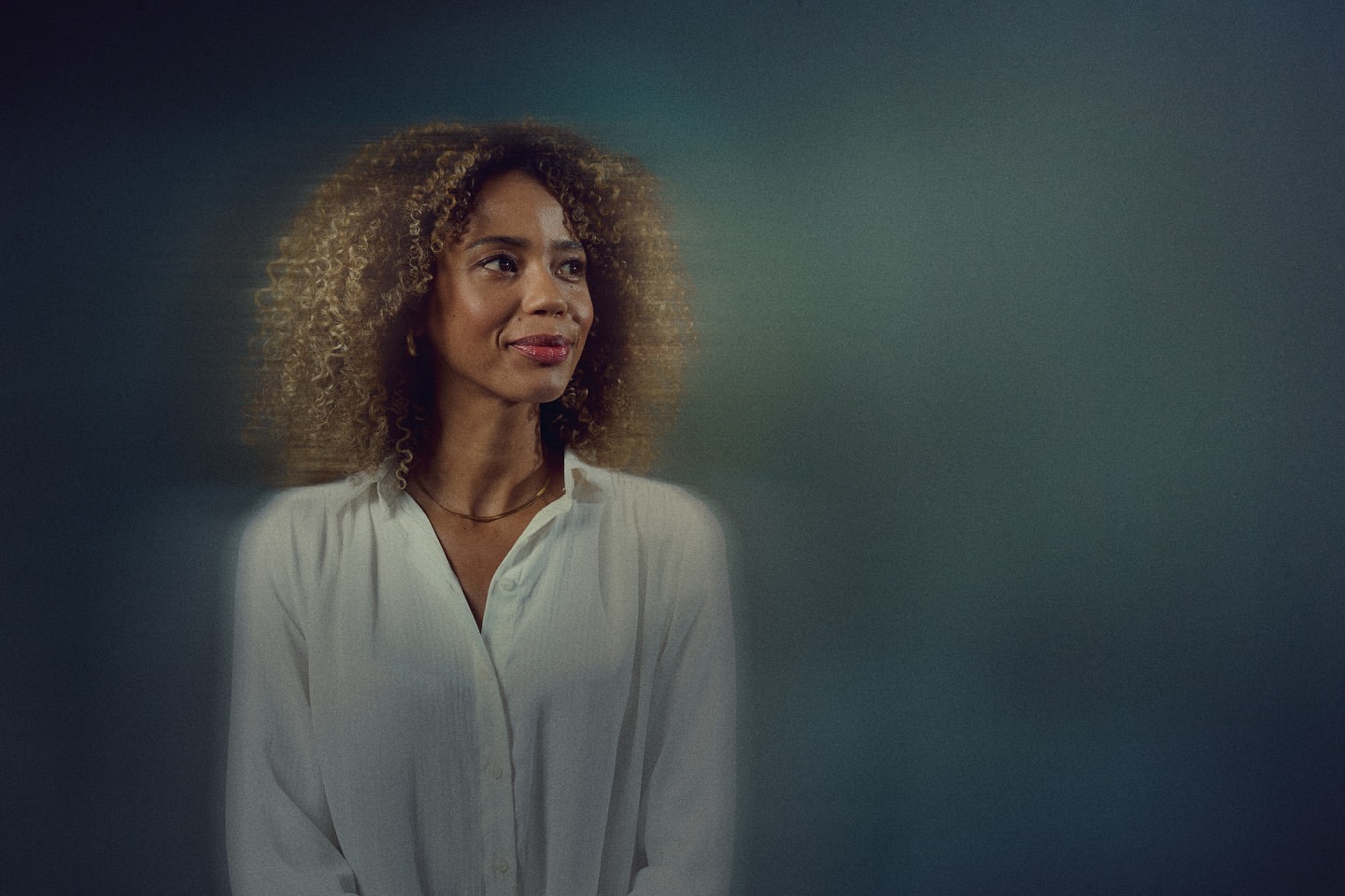 Portrait of a smiling African-American woman with curly hair wearing a white blouse against a dark teal and blue gradient background.