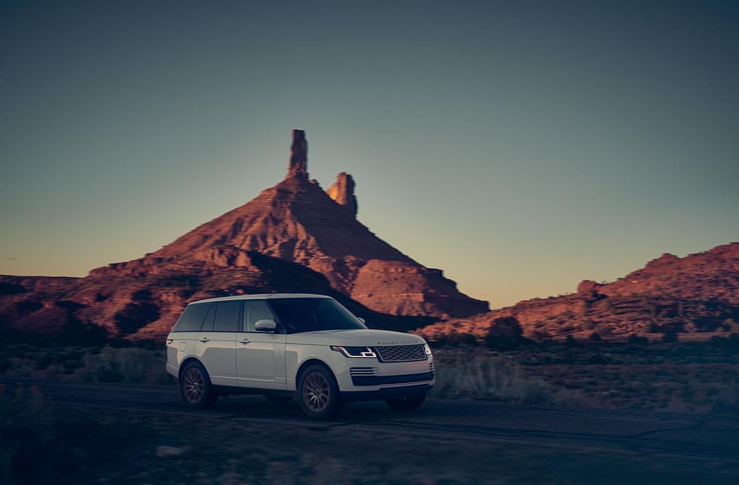 White Range Rover SUV driving on a dirt road with a dramatic mesa rock formation in the background under a twilight sky.