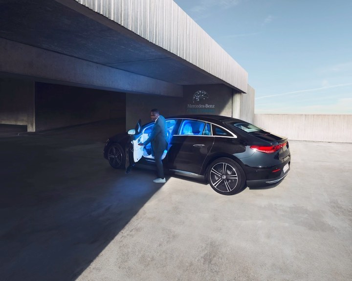 An African American man exits a black Mercedes-Benz EQS sedan in an urban parking structure with concrete walls and blue interior lighting.