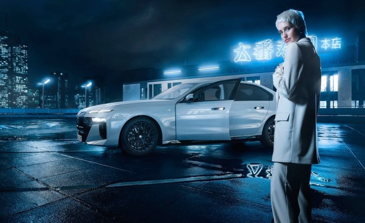 Fashionable woman in a suit standing beside a silver sedan car under a neon sign in a nighttime city scene. Automotive promotional shot.