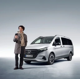 Man holding a smartphone standing beside a silver electric van in a studio setting