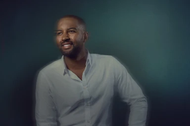 Portrait of a smiling Black man in a white collared shirt against a dark green textured background with motion blur effect.