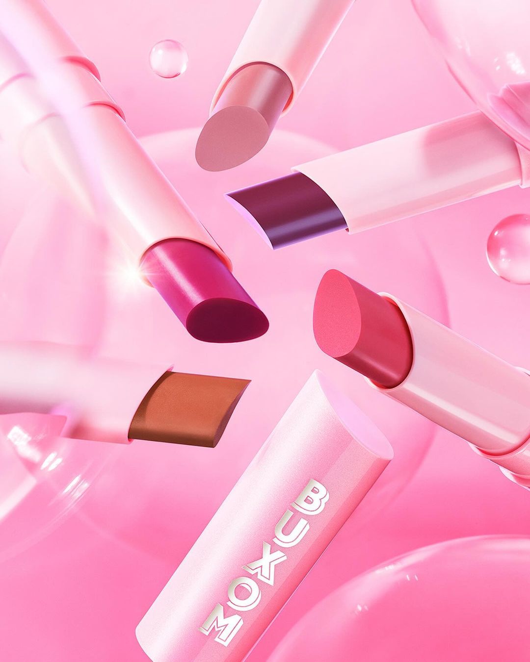 Floating Buxom lipsticks on a soft pink background with translucent spheres create a beauty product advertisement featuring various shades.