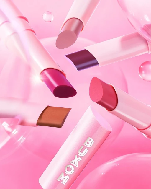 Floating Buxom lipsticks on a soft pink background with translucent spheres create a beauty product advertisement featuring various shades.