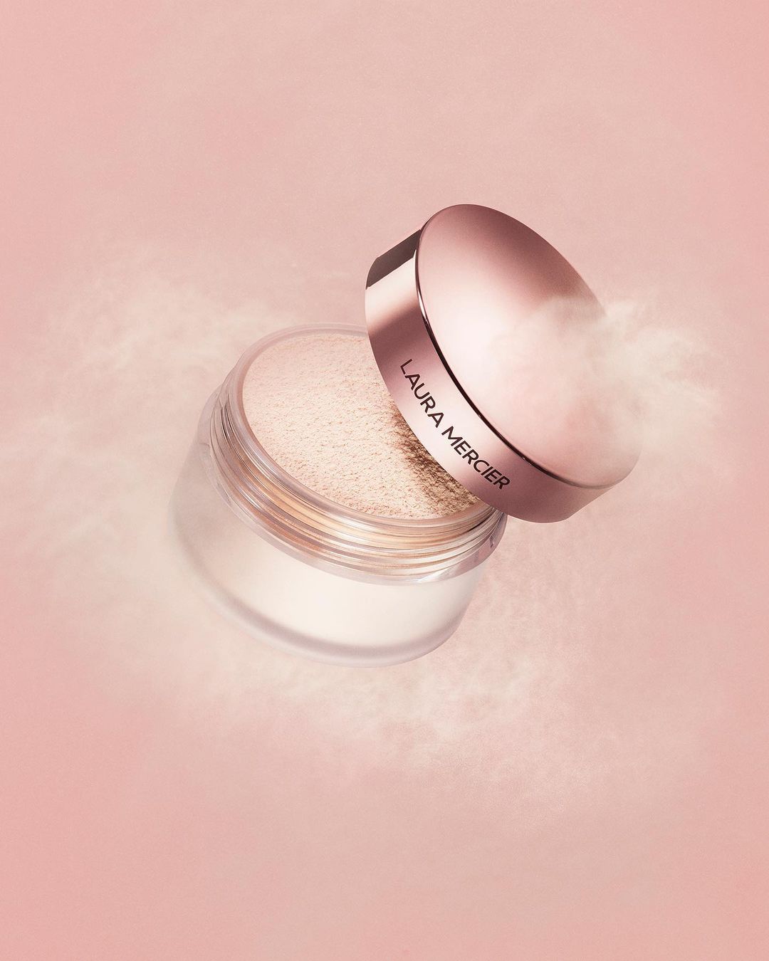 Close-up studio shot of Laura Mercier translucent loose setting powder jar with lid askew, on light pink backdrop with hazy powder effect.