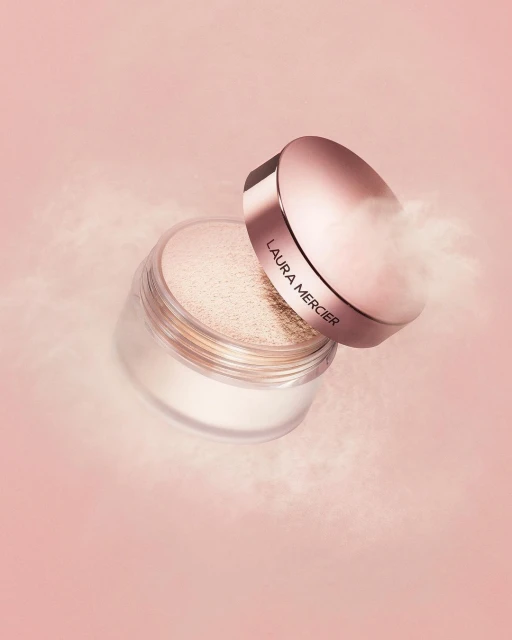 Close-up studio shot of Laura Mercier translucent loose setting powder jar with lid askew, on light pink backdrop with hazy powder effect.