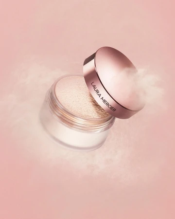 Close-up studio shot of Laura Mercier translucent loose setting powder jar with lid askew, on light pink backdrop with hazy powder effect.