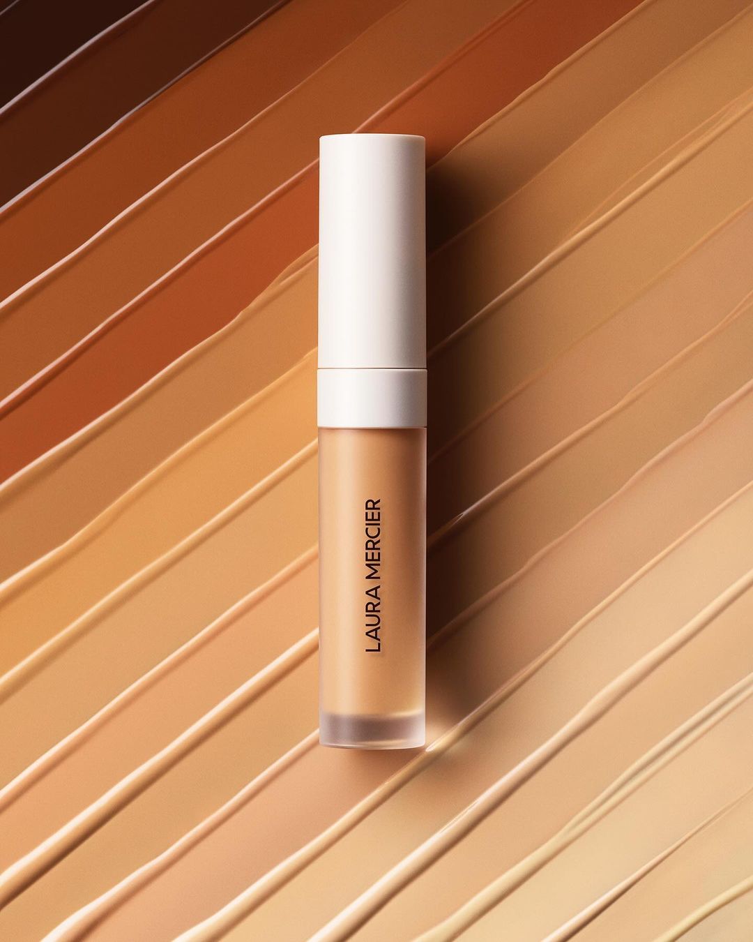 Laura Mercier concealer wand displayed diagonally against a background of diagonal strokes of multiple shades of tan foundation.