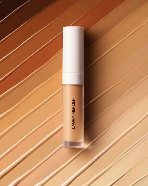 Laura Mercier concealer wand displayed diagonally against a background of diagonal strokes of multiple shades of tan foundation.