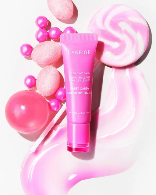 Pink Laneige lip glowy balm tube surrounded by lollipops and pink candies. Beauty product shot, makeup promotion, pink aesthetic.