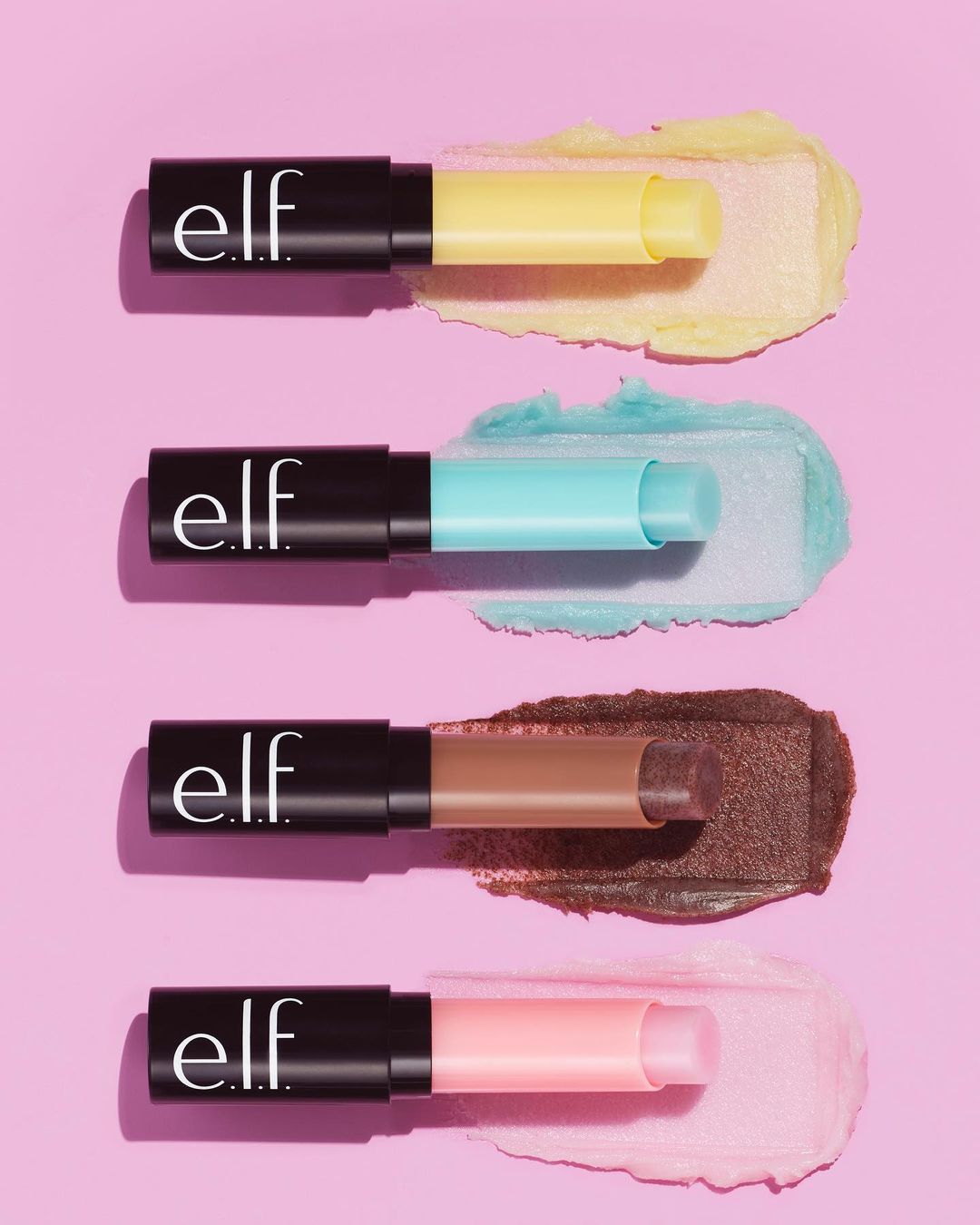 Four e.l.f. lip exfoliator sticks in various pastel shades (yellow, blue, brown, pink) with matching scrub swatches on a pink background.