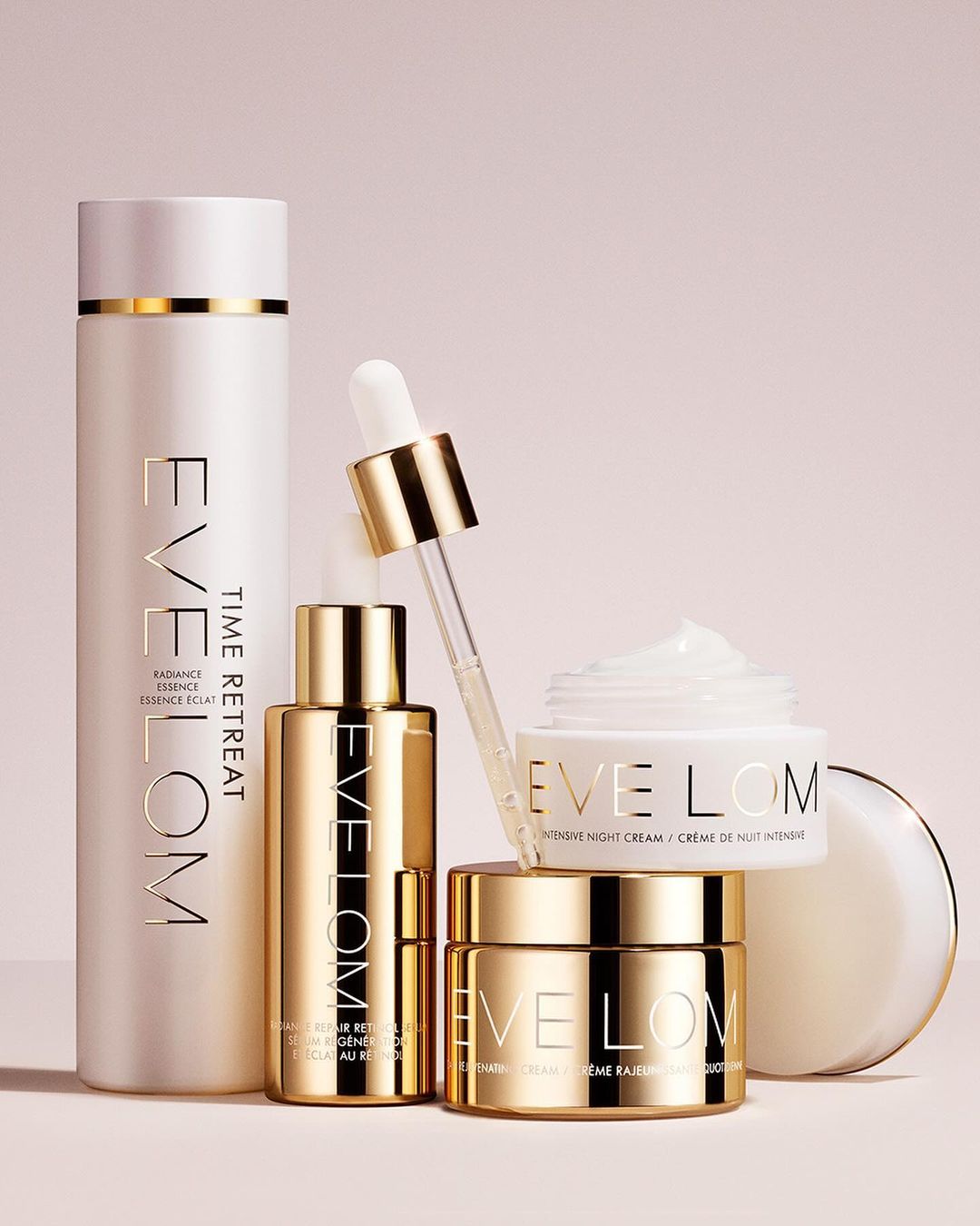 Arrangement of Eve Lom skincare products in gold and white packaging including cleanser, serum, and night cream against a pale pink background.