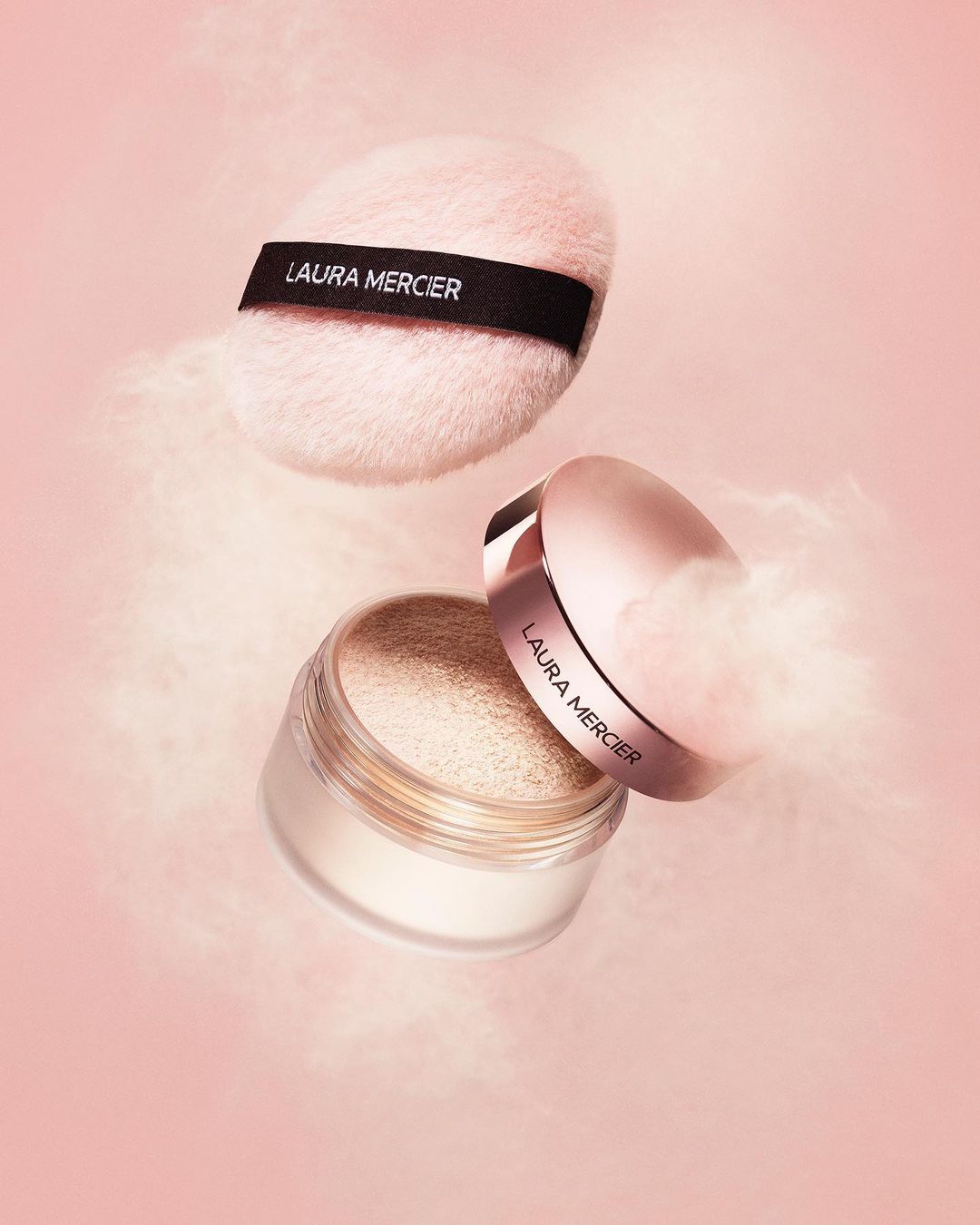 Laura Mercier powder and puff on pink background. Powder floats around the makeup products in a beauty product promotional shot.