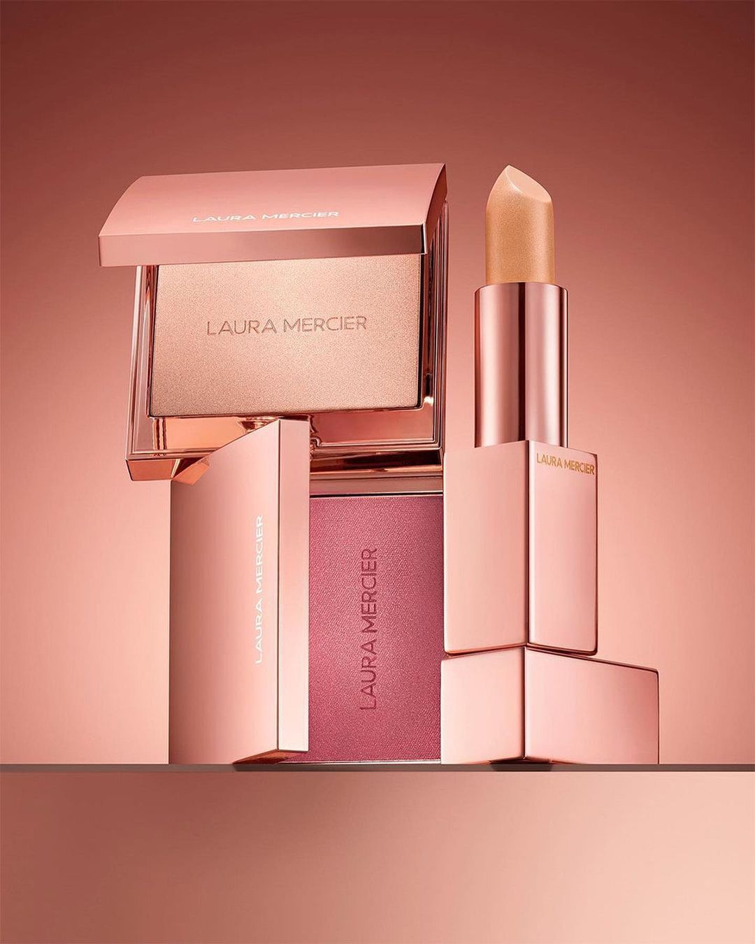 Laura Mercier makeup products in rose gold packaging include lipstick, blush and highlighting powder, arranged on a reflective surface.