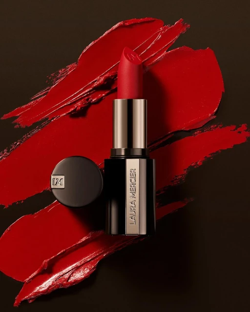 Laura Mercier lipstick with lid on black background. Red lipstick is in the foreground with smears of red lipstick in the background.