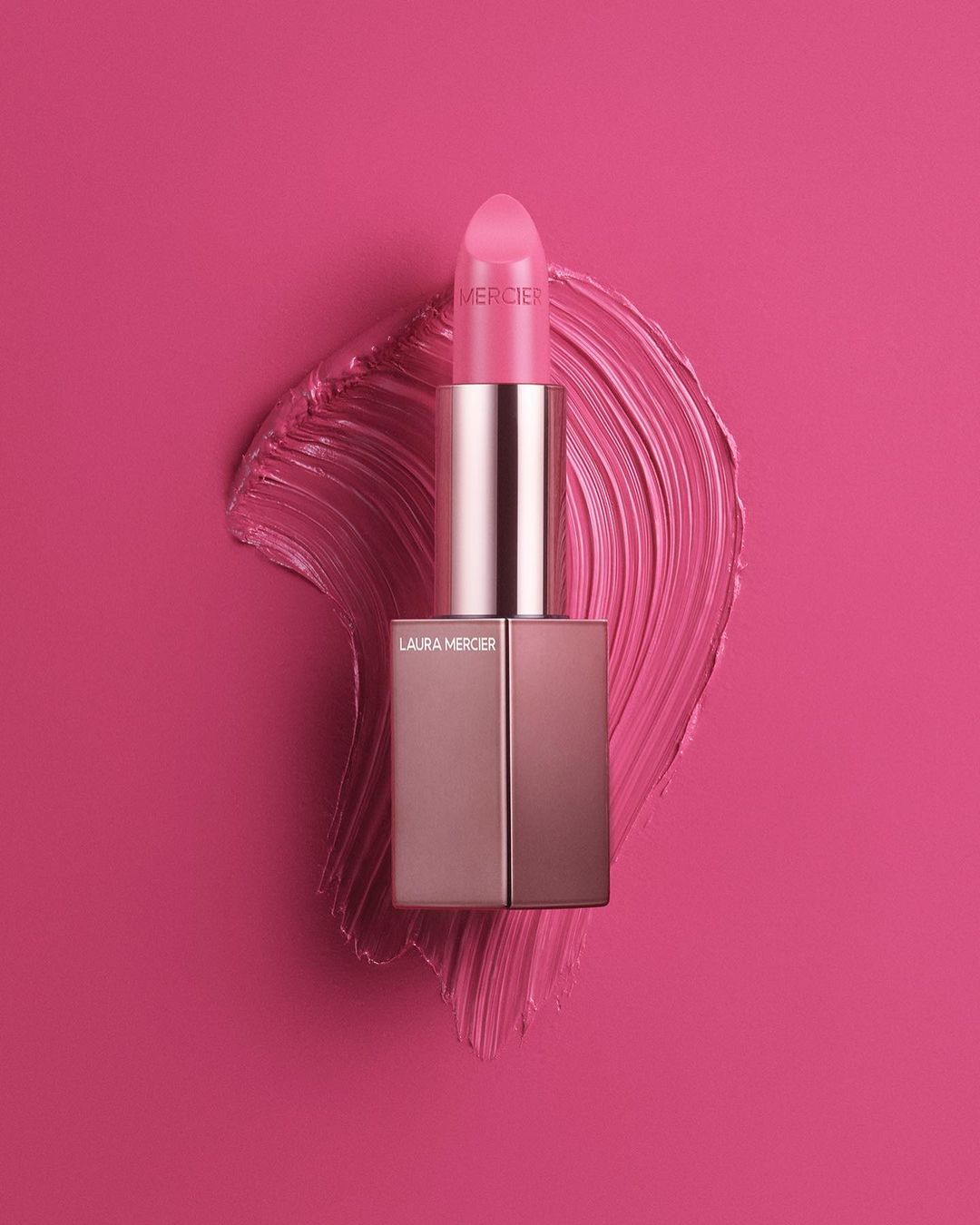 Close-up studio shot of Laura Mercier lipstick in pink with a smear of matching lipstick against a vivid pink backdrop.