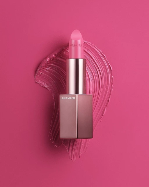 Close-up studio shot of Laura Mercier lipstick in pink with a smear of matching lipstick against a vivid pink backdrop.