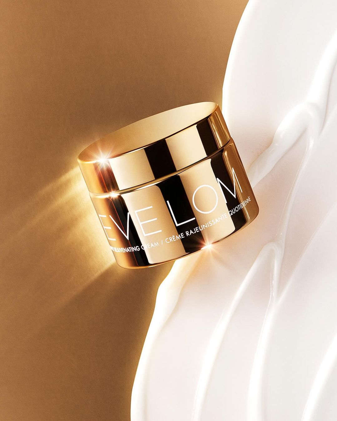 Close-up studio shot of a gold jar of Eve Lom Rejuvenating Cream set against a swirl of white cream and a beige background.