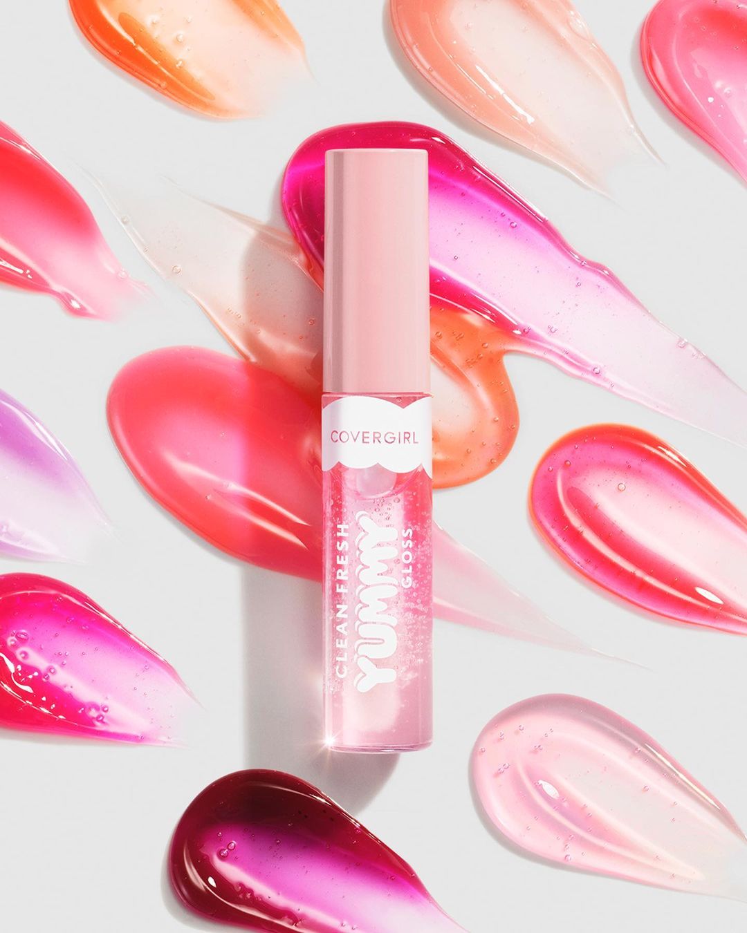 A tube of CoverGirl lip gloss surrounded by swipes of shimmery gloss in various shades of pink, peach, and berry tones.