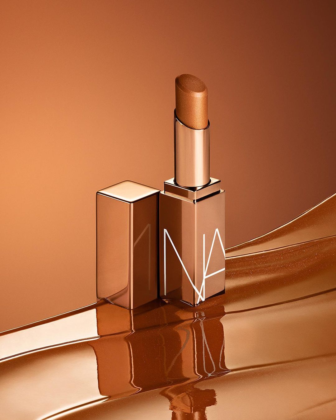 Close-up studio shot of a NARS lipstick with the lid off on a reflective bronze surface against a bronze gradient background.