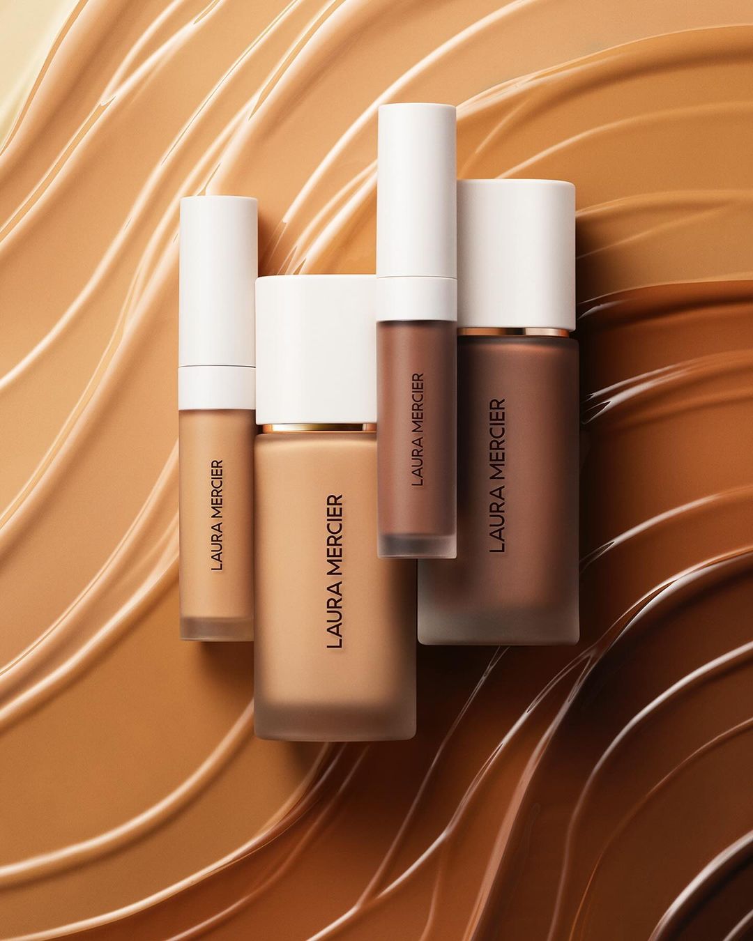 Laura Mercier brand makeup product tubes and bottles of foundation in neutral color palette on a light brown and tan swirled background.