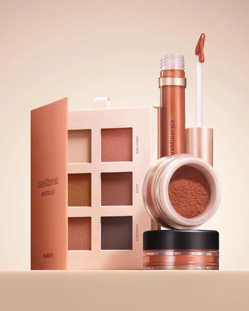 BareMinerals makeup products including eyeshadow palettes, powder and lip gloss are displayed on a beige surface against a neutral background.