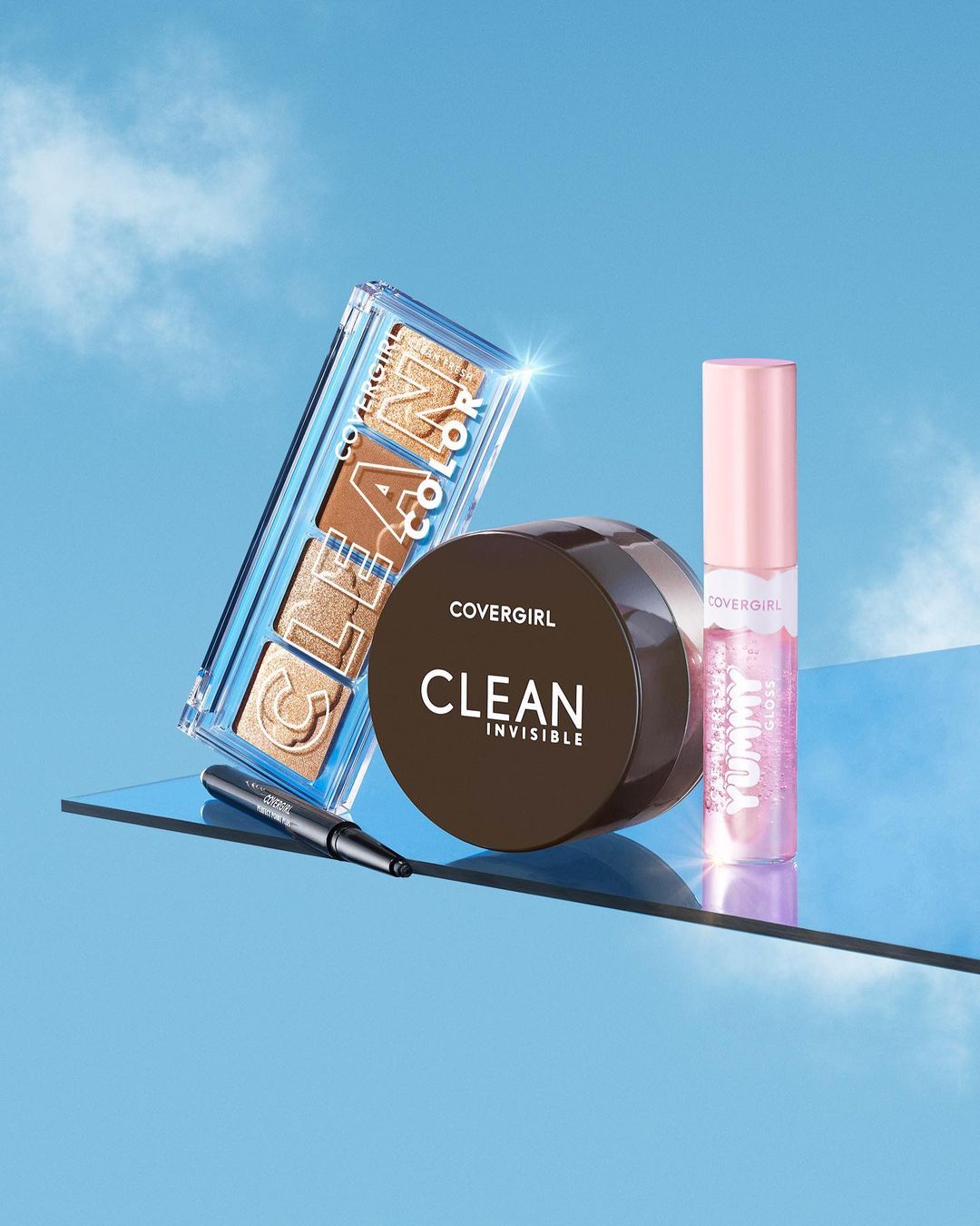 Still life shot of COVERGIRL makeup: an eye palette, a jar of foundation, an eyeliner, and a lip gloss against a blue sky.