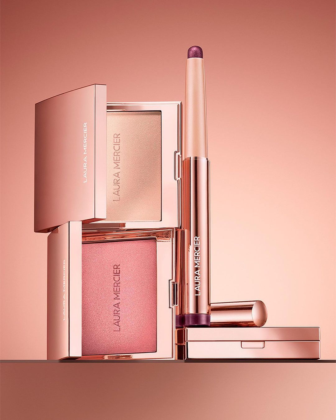 Cosmetic product still life: rose gold Laura Mercier compacts and stick arranged on a matching surface, warm lighting, beauty concept.